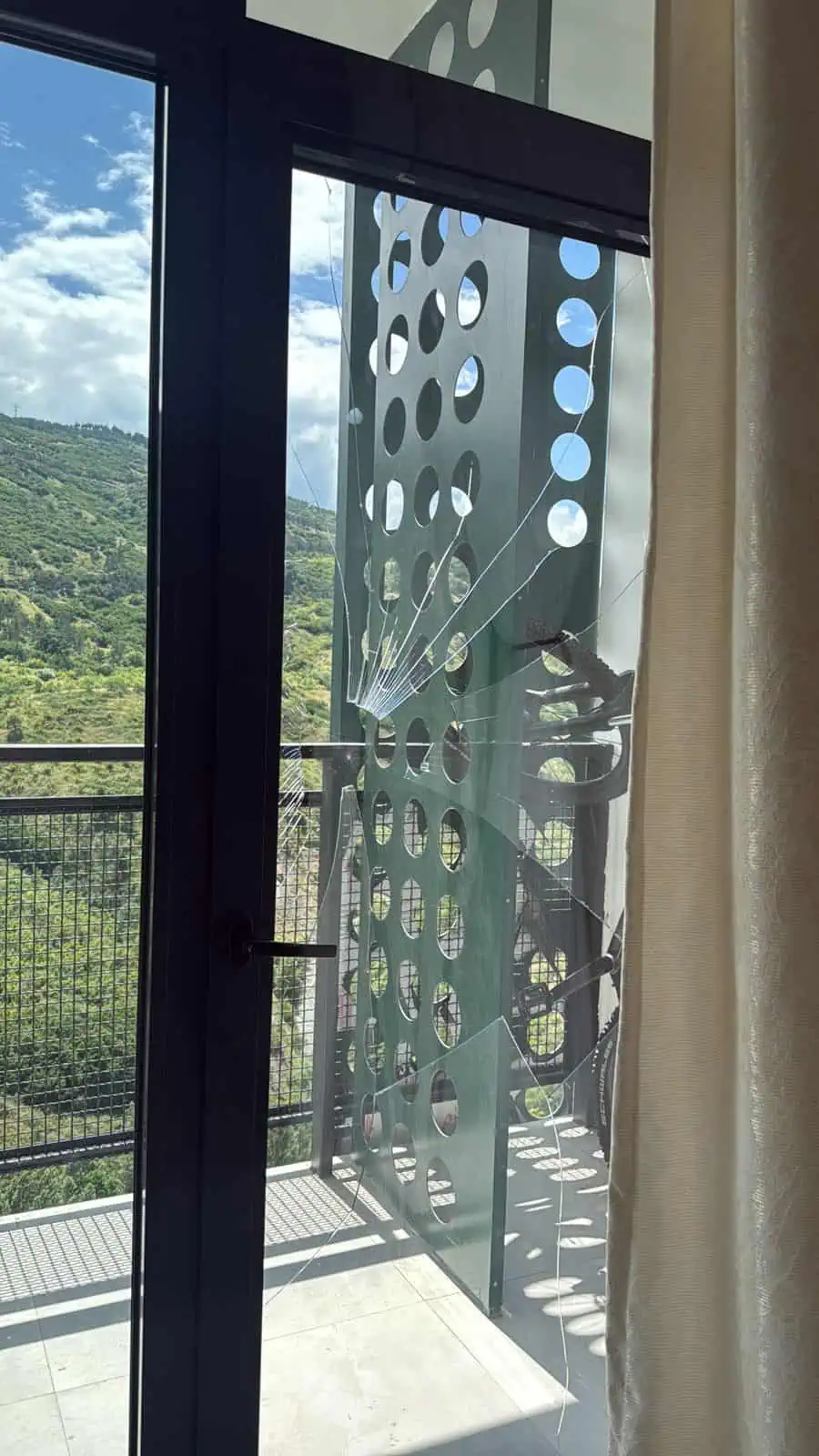 Cracked balcony door glazing unit with spider-web fractures on a green mountain backdrop in Tbilisi